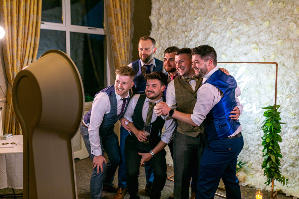 Groomsmen in waistcoats and ties laughing together at a wedding photo booth in front of a white flower wall with a neon sign.