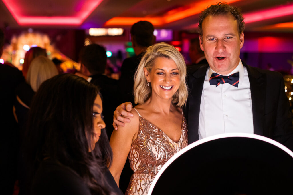 Smiling couple in formal evening wear reacting to their photo at a ring-light photo booth during a glamorous gala event.